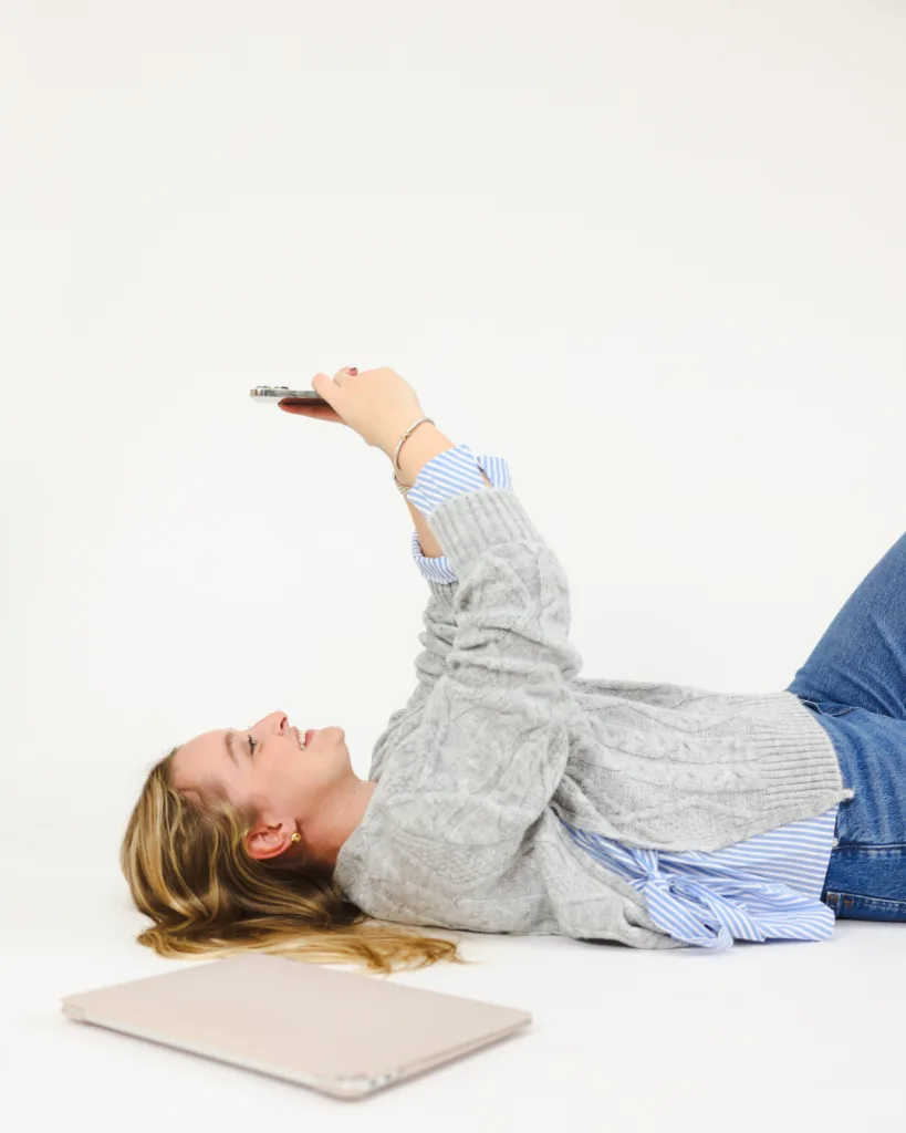 woman lays next to a laptop while working on a cell phone
